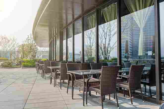 Outdoor cafe renovated in Romford with wicker tables and chairs, sunlight filtering through large glass windows, surrounded by greenery and city skyline.