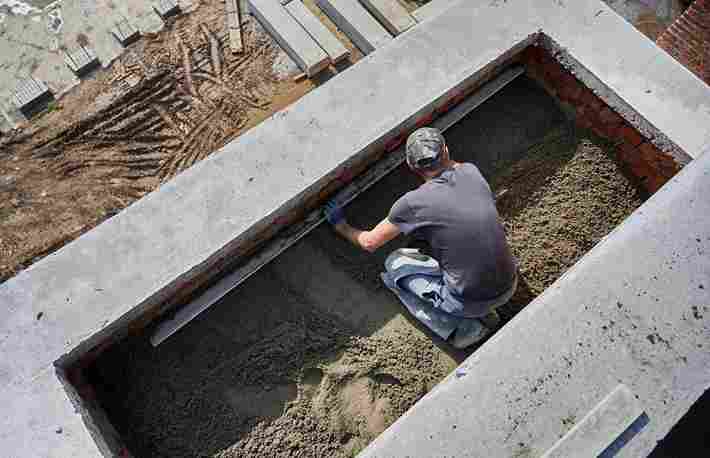 A worker in a cap levels concrete in Romford in a construction site trench, surrounded by tools and materials.