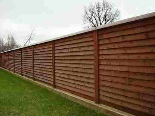 A long wooden fence with horizontal slats installed in Romford runs parallel to a grassy area, under a cloudy sky, with sparse trees in the background.
