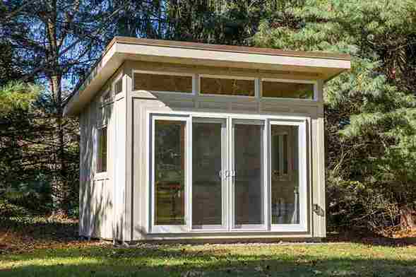 A modern, small garden shed built in Romford with large glass doors and windows, nestled among trees in a lush green yard.