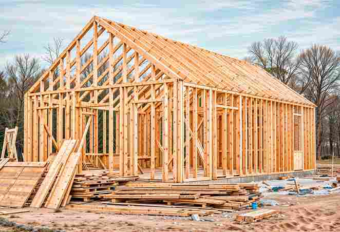 A wooden frame house under construction, surrounded by stacks of lumber and tools, against a backdrop of trees and cloudy sky.