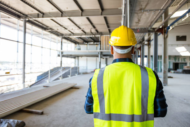 A person in a yellow hard hat and reflective vest stands inside a large, unfinished building with exposed beams and windows.