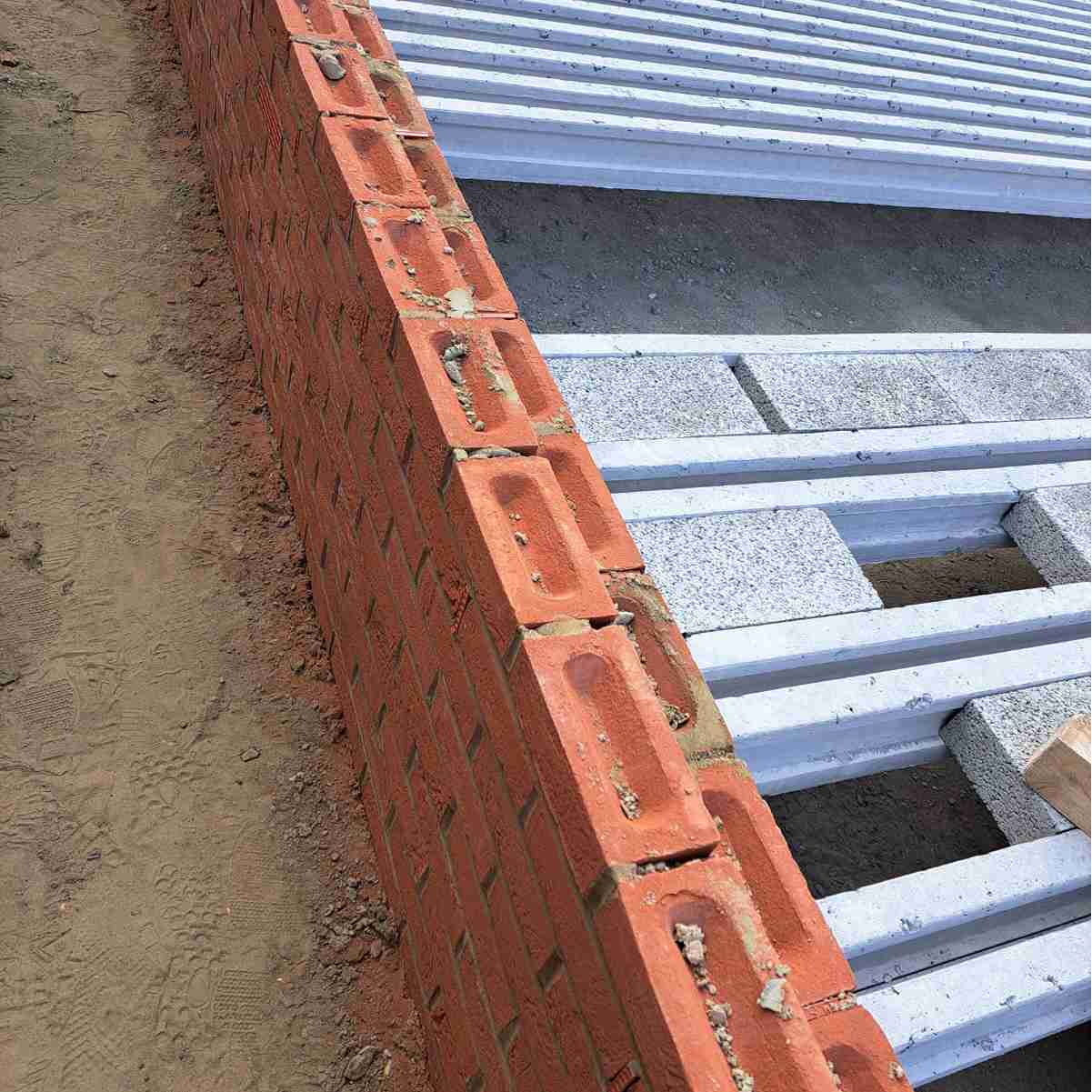 A close-up of a partially constructed red brick wall with mortar, adjacent to stacked metal beams and gravel on a construction site.