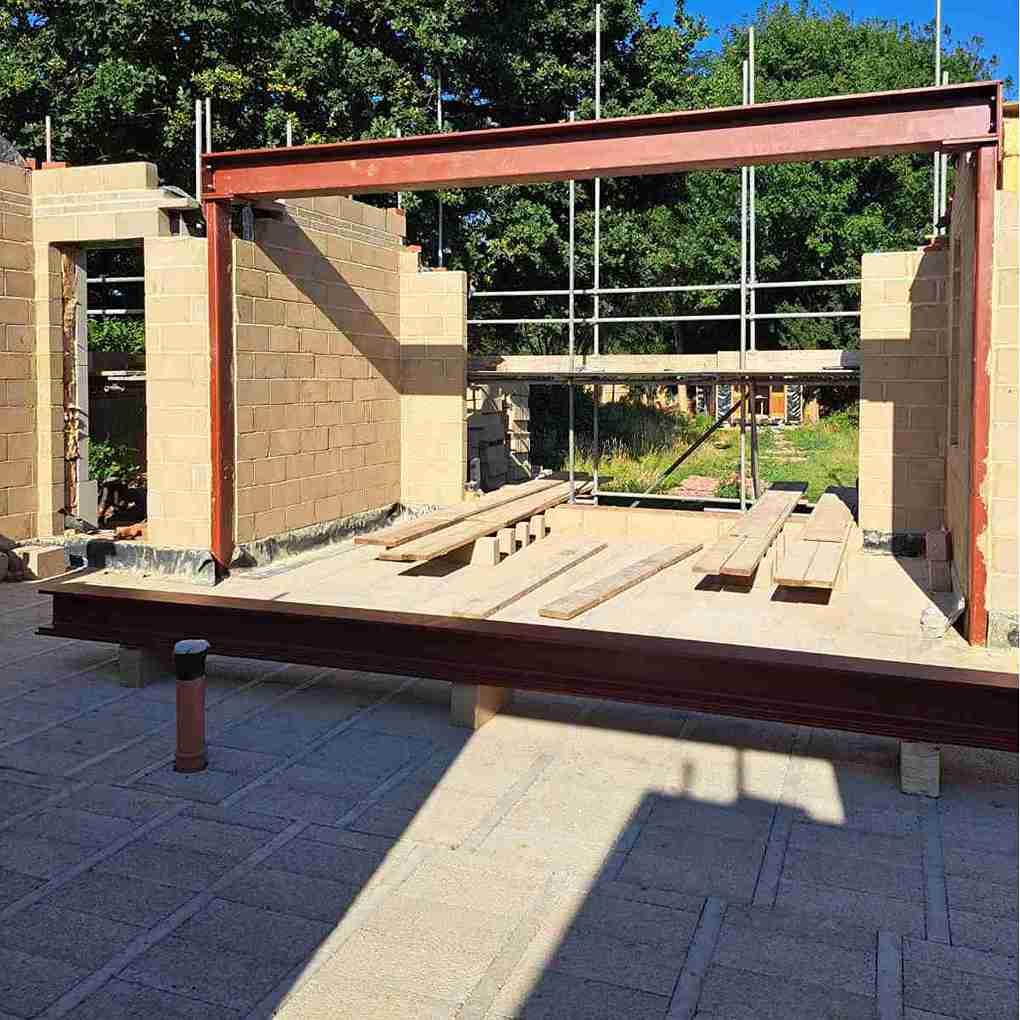 Construction site featuring a steel beam and concrete block walls, with scaffolding and wooden planks on a paved floor.