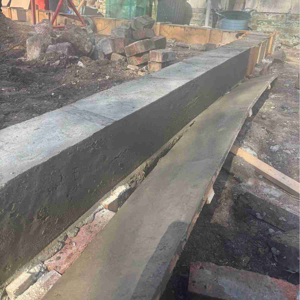 A freshly poured concrete beam and pathway, surrounded by stacked bricks and dirt, in a construction site setting.