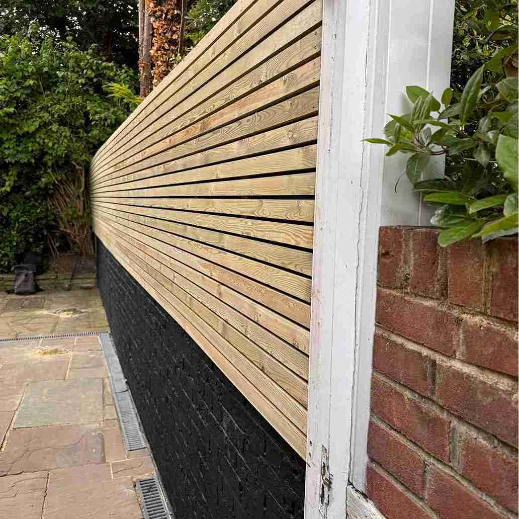 A wooden slatted fence runs alongside a painted black brick wall, with greenery and a brick border visible in the background.