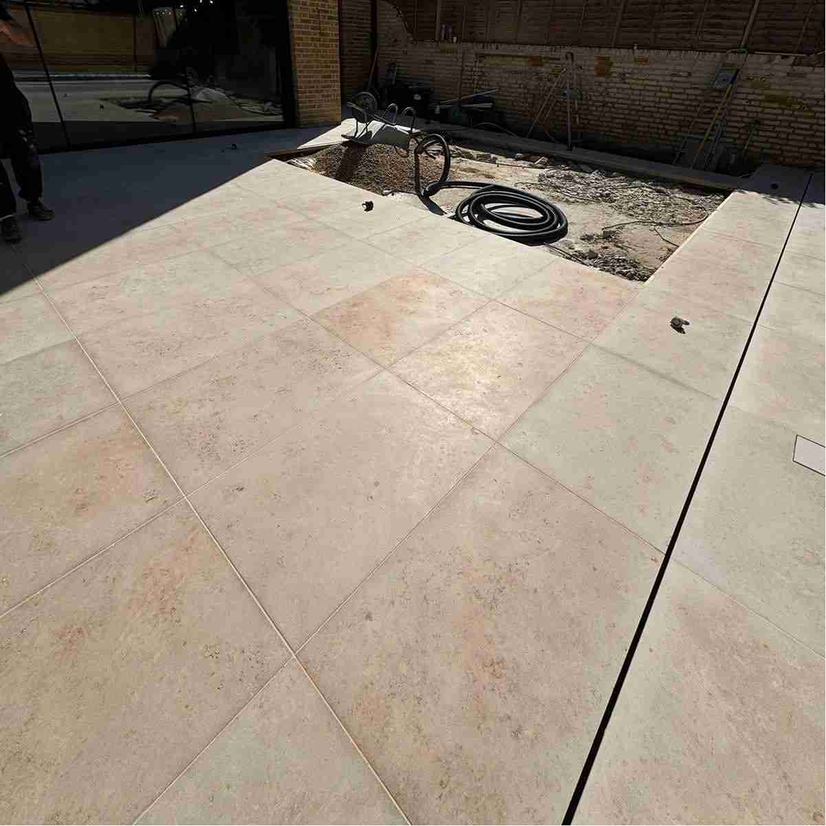 A sunlit patio with large light-colored tiles, featuring a partially excavated area and construction materials in the background.