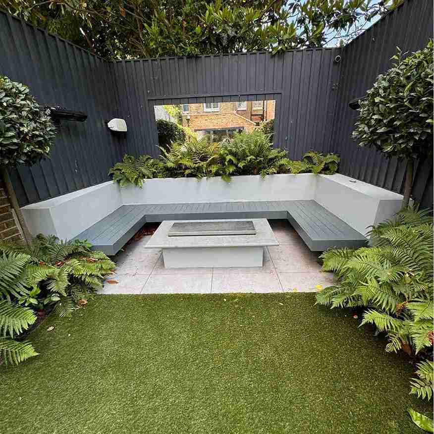 Cozy outdoor seating area with gray benches, a central fire pit, and lush ferns, enclosed by dark fencing and greenery.