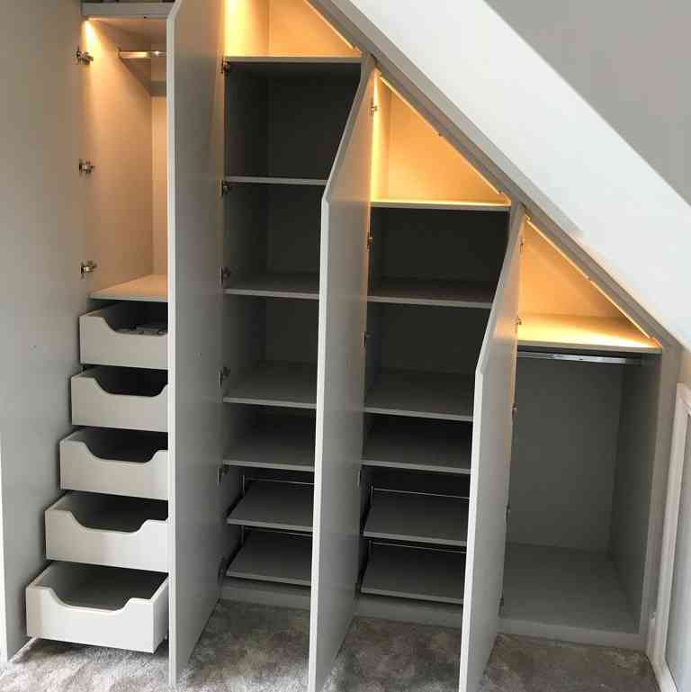A sleek, modern closet under a staircase featuring illuminated shelves, drawers, and organized storage spaces in light gray.