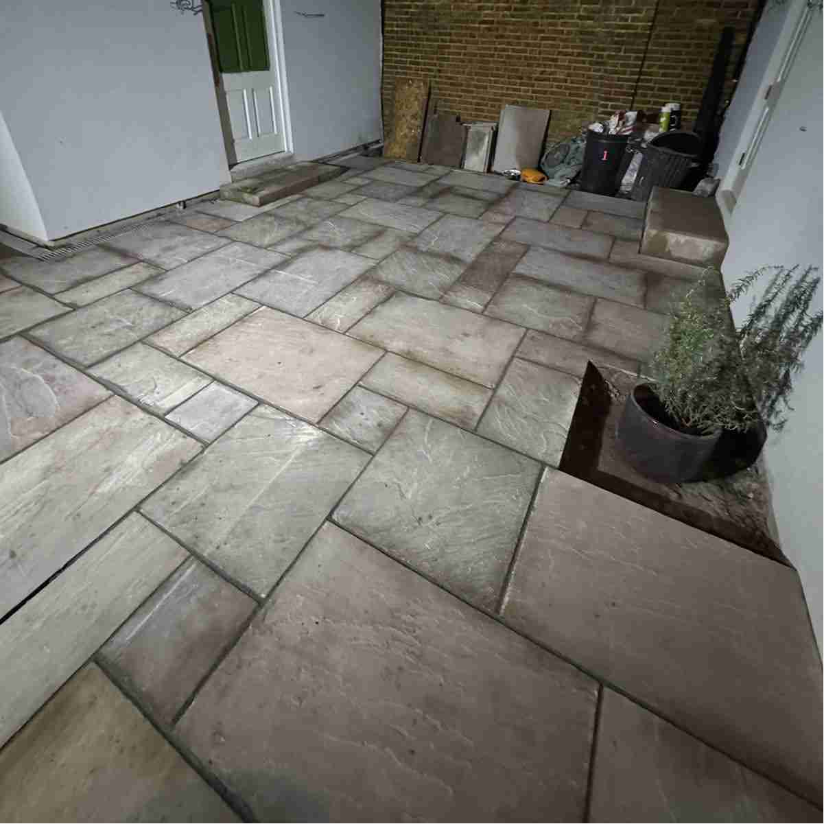 A newly laid stone patio featuring irregular slabs in muted tones, with potted rosemary and construction materials in the background.