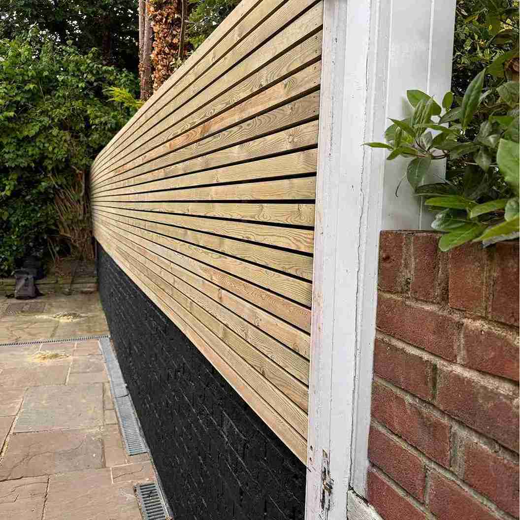 Wooden slatted fence installation alongside a black-painted brick wall, with greenery in a residential garden in East London.
