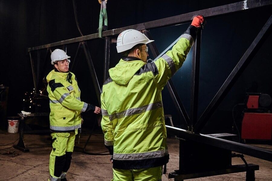 Two workers in high-visibility jackets and helmets operate a metallic structure in a dimly lit industrial setting.